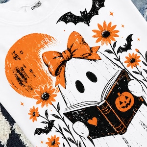 Pu&ograve; includere: Una t-shirt bianca con una stampa grafica di un fantasma che indossa un fiocco arancione, intento a leggere un libro nero con l'illustrazione di una zucca. Il design include una luna arancione, pipistrelli neri e fiori arancioni.