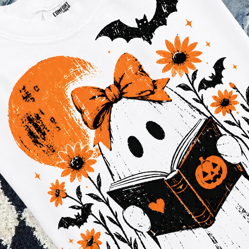 Pu&ograve; includere: Una t-shirt bianca con una stampa grafica di un fantasma che indossa un fiocco arancione, intento a leggere un libro nero con l'illustrazione di una zucca. Il design include una luna arancione, pipistrelli neri e fiori arancioni.