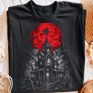May include: A black t-shirt featuring a graphic print of a dark, multi-story haunted house in a graveyard. Above the house is a large, vibrant red full moon with a silhouette of a witch flying on a broomstick and several bats in flight.