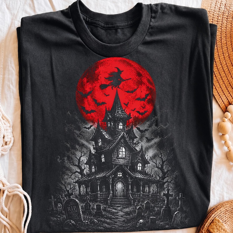 May include: A black t-shirt featuring a graphic print of a dark, multi-story haunted house in a graveyard. Above the house is a large, vibrant red full moon with a silhouette of a witch flying on a broomstick and several bats in flight.