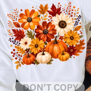 Puede incluir: Una camiseta blanca con un estampado gráfico vibrante de elementos otoñales, que incluye flores naranjas, amarillas y rojas, varias hojas de otoño, pequeñas bayas y tres calabazas en tonos naranja y blanco dispuestas en un grupo.