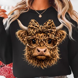 Pode incluir: Camisola preta com um design de vaca das Terras Altas. A vaca tem um la&ccedil;o com estampa de leopardo, &oacute;culos e pelo. A imagem &eacute; um close-up do rosto da vaca, com chifres e um nariz rosa. O design est&aacute; centrado.