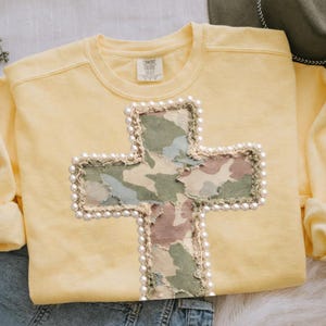 Pode incluir: Um moletom amarelo Comfort Colors com um grande design de cruz. A cruz é feita de tecido camuflado e é contornada com pérolas falsas. O moletom tem mangas compridas e gola redonda.