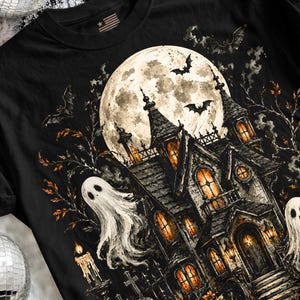 May include: A black t-shirt featuring a graphic print of a haunted house under a large full moon. The design includes two white ghosts, flying bats, bare tree branches, and a lit candle. The windows of the house glow with a warm orange light.
