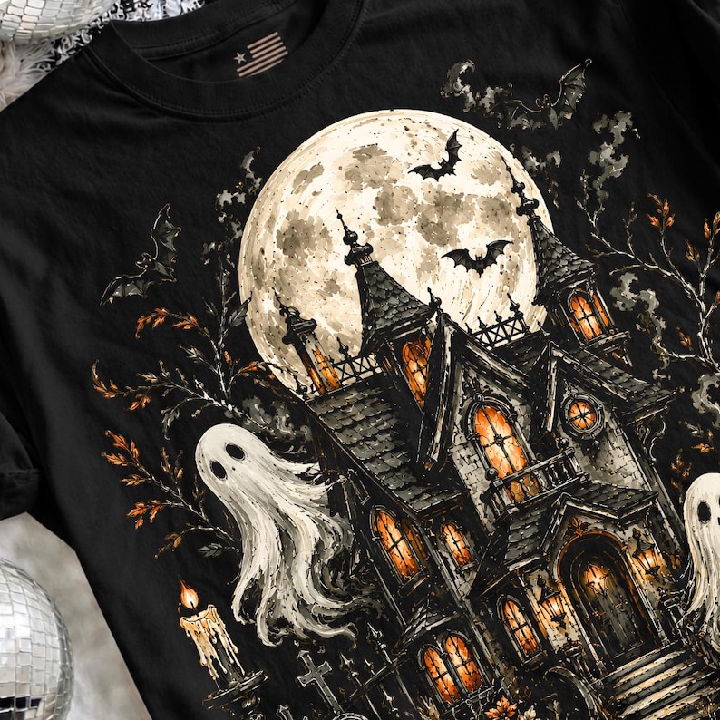 May include: A black t-shirt featuring a graphic print of a haunted house under a large full moon. The design includes two white ghosts, flying bats, bare tree branches, and a lit candle. The windows of the house glow with a warm orange light.