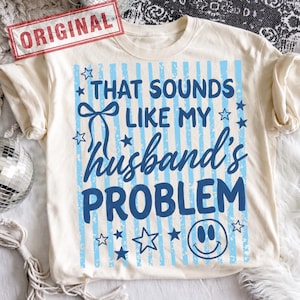 May include: Cream-colored t-shirt with the text "THAT SOUNDS LIKE MY husband's PROBLEM" in blue. The design includes blue vertical stripes, stars, and a smiley face. The word "ORIGINAL" is stamped in red.