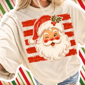 May include: A cream-colored t-shirt featuring a graphic of a winking Santa Claus face with a red hat, white beard, and holly berries. The design is layered over horizontal red brushstroke stripes, creating a festive holiday-themed graphic print.