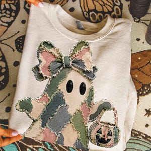 Könnte beinhalten: Ein beigefarbenes Sweatshirt mit Rundhalsausschnitt und einer großen Applikation eines Geistes mit Schleife. Der Geist besteht aus ausgefransten, mehrfarbigen Stoffstücken in Rosa-, Grün- und Blautönen und hält einen kleinen Kürbiseimer.
