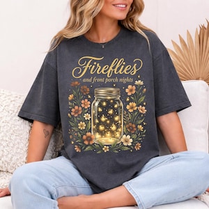 Pu&ograve; includere: T-shirt grigio scuro con un barattolo pieno di lucciole luminose, circondato da fiori arancioni, gialli e bianchi. Il testo "Fireflies and front porch nights" &egrave; scritto in oro.