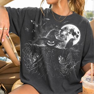 May include: A charcoal gray short-sleeve t-shirt featuring a graphic print of a scarecrow with a glowing jack-o'-lantern head, wearing a wide-brimmed hat. The design includes a large full moon, a perched raven, and flying bats in white.