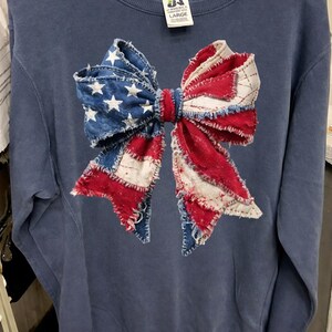 K&ouml;nnte beinhalten: Marineblaues Sweatshirt mit einem gro&szlig;en Schleifendesign. Die Schleife besteht aus Denim sowie rotem und wei&szlig;em Stoff, der an die amerikanische Flagge erinnert. Das Sweatshirt hat einen Rundhalsausschnitt und lange &Auml;rmel. Das Etikett lautet "LARGE".