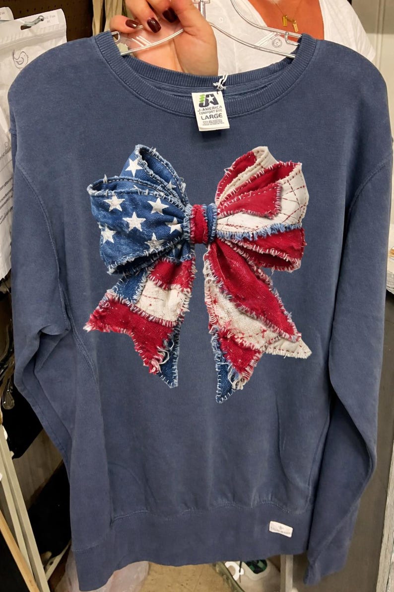 K&ouml;nnte beinhalten: Marineblaues Sweatshirt mit einem gro&szlig;en Schleifendesign. Die Schleife besteht aus Denim sowie rotem und wei&szlig;em Stoff, der an die amerikanische Flagge erinnert. Das Sweatshirt hat einen Rundhalsausschnitt und lange &Auml;rmel. Das Etikett lautet "LARGE".