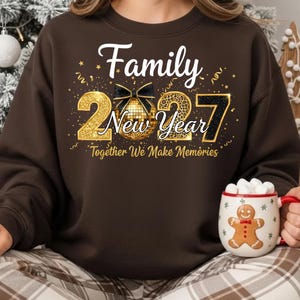 May include: A dark brown crewneck sweatshirt featuring a graphic with the text Family New Year 2027 Together We Make Memories. The design includes a gold disco ball with a black bow, gold glitter numbers, and gold confetti accents on the front.