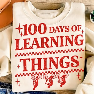 May include: Cream-coloured sweatshirt featuring the red text "100 Days of Learning Things". The design includes bicycle illustrations and star accents, ideal for celebrating an educational milestone.