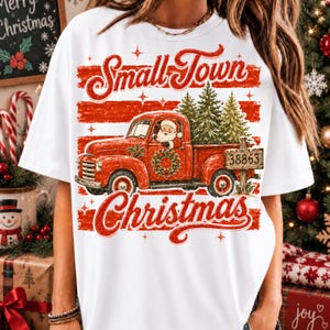 May include: A white t-shirt featuring a graphic of a red vintage pickup truck carrying green pine trees. Santa Claus is in the driver's seat. The design includes a wreath on the door, the text 38863, and the words Small Town Christmas in red.