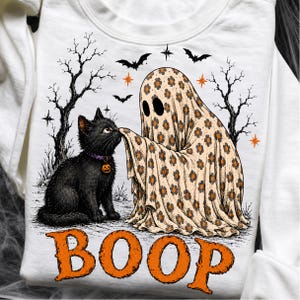 May include: A white sweatshirt featuring a graphic of a black cat with a purple collar and a pumpkin charm, touching noses with a ghost covered in a beige sheet with a brown flower pattern. The design includes black trees, bats, and the word BOOP.
