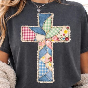 May include: Dark grey t-shirt with a patchwork cross design. The cross is made from denim, gingham, and floral fabrics, with a cream border. The shirt has short sleeves and a crew neck. The measurements are in centimetres.
