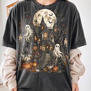 May include: A charcoal gray t-shirt featuring a detailed graphic print of a haunted mansion under a full moon. The design includes two white ghosts, orange pumpkins, lit candles, bare trees, and flying bats, creating a dark, atmospheric scene.