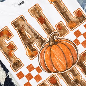 May include: A white t-shirt featuring the word FALL in large block letters filled with a brown camouflage and orange maple leaf pattern. An orange pumpkin graphic is positioned over the letters, and orange and white checkered squares are included.