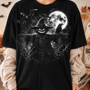 May include: A black t-shirt featuring a graphic print of a scarecrow with a glowing jack-o'-lantern head, wearing a pointed hat. The scene includes a large full moon, a perched raven, and flying bats, all rendered in a high-contrast black and white style.