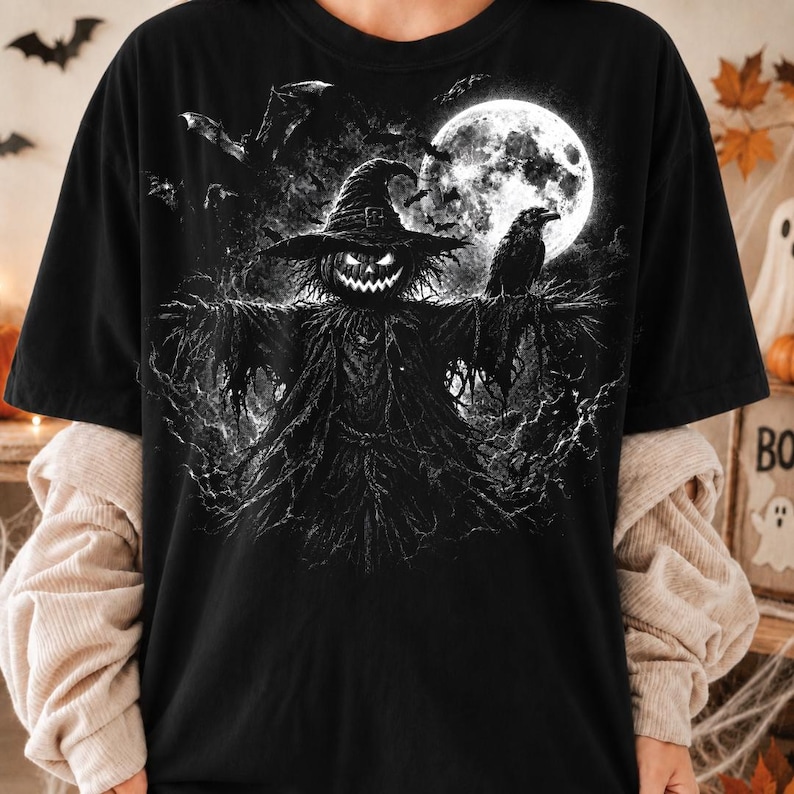 May include: A black t-shirt featuring a graphic print of a scarecrow with a glowing jack-o'-lantern head, wearing a pointed hat. The scene includes a large full moon, a perched raven, and flying bats, all rendered in a high-contrast black and white style.