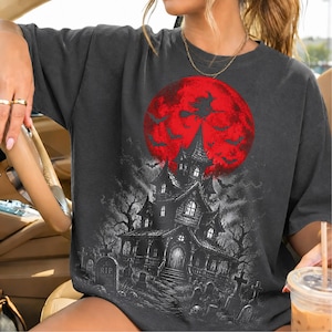 May include: A charcoal gray t-shirt featuring a graphic print of a haunted house in a graveyard under a large red moon. The design includes silhouettes of bats and a witch flying on a broomstick against the moon, with detailed tombstones.