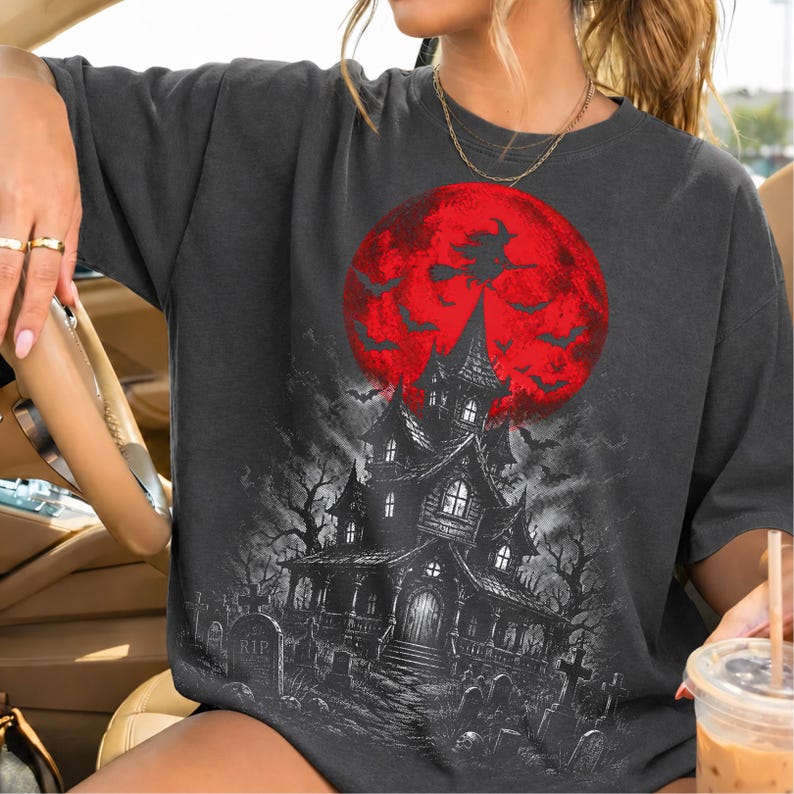 May include: A charcoal gray t-shirt featuring a graphic print of a haunted house in a graveyard under a large red moon. The design includes silhouettes of bats and a witch flying on a broomstick against the moon, with detailed tombstones.