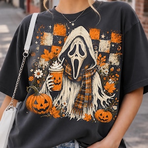 May include: A charcoal gray t-shirt featuring a graphic of a hooded ghost figure with a mask, holding a pumpkin-faced cup. The design includes orange and black checkered patterns, autumn leaves, daisies, and two jack-o'-lanterns at the base.