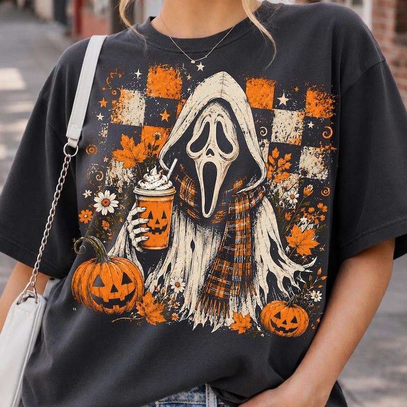 May include: A charcoal gray t-shirt featuring a graphic of a hooded ghost figure with a mask, holding a pumpkin-faced cup. The design includes orange and black checkered patterns, autumn leaves, daisies, and two jack-o'-lanterns at the base.