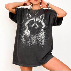 Pu&ograve; includere: T-shirt grigio scuro con una grafica bianca di un procione con occhiali da sole che soffia una bolla. Il procione fa il dito medio. La maglietta ha una vestibilit&agrave; ampia e maniche corte.