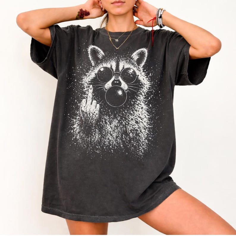 Pu&ograve; includere: T-shirt grigio scuro con una grafica bianca di un procione con occhiali da sole che soffia una bolla. Il procione fa il dito medio. La maglietta ha una vestibilit&agrave; ampia e maniche corte.