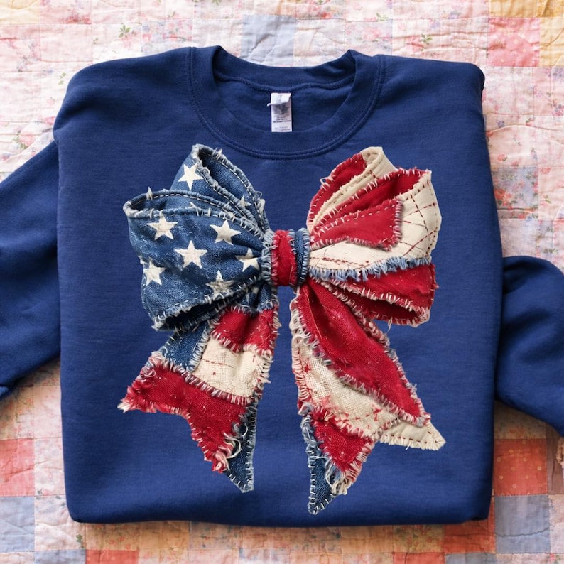 K&ouml;nnte beinhalten: Ein marineblaues Sweatshirt mit einem gro&szlig;en Schleifendesign. Die Schleife besteht aus Denim sowie rotem und wei&szlig;em Stoff mit einem Used-Patchwork-Look. Die linke Seite der Schleife hat ein blaues Denim-Muster mit wei&szlig;en Sternen.