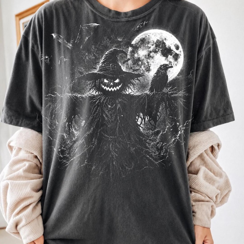May include: A charcoal gray short-sleeve t-shirt featuring a graphic print of a scarecrow with a glowing jack-o'-lantern head, wearing a wide-brimmed hat. A raven sits on its shoulder against a backdrop of a large full moon and flying bats.