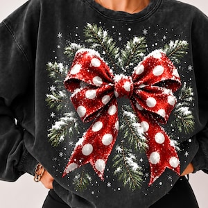 Peut inclure: Un sweat-shirt à col rond gris anthracite orné d'un grand motif central représentant un nœud rouge à pois blancs. Le nœud est superposé sur des branches de pin vertes saupoudrées de neige blanche, avec de petits flocons blancs dispersés.