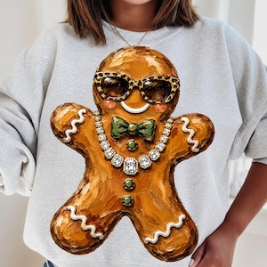 Könnte beinhalten: Ein hellgraues Sweatshirt mit Rundhalsausschnitt und einer großen, strukturierten, malerischen Grafik eines Lebkuchenmanns. Die Lebkuchenfigur trägt eine Sonnenbrille mit Leopardenmuster, eine grüne Fliege, eine Kristallkette und zwei grüne Knöpfe.