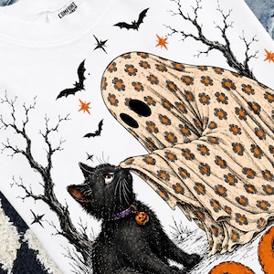 May include: A white sweatshirt featuring a graphic of a ghost covered in an orange and black floral pattern interacting with a black cat wearing a purple collar with a pumpkin charm, surrounded by black bats, bare trees, and orange stars.