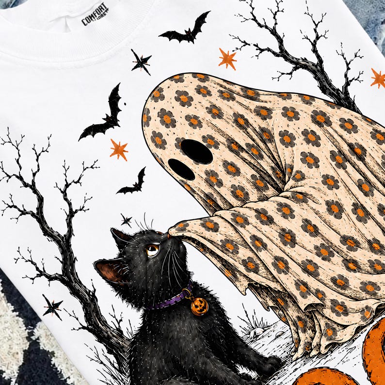 May include: A white sweatshirt featuring a graphic of a ghost covered in an orange and black floral pattern interacting with a black cat wearing a purple collar with a pumpkin charm, surrounded by black bats, bare trees, and orange stars.