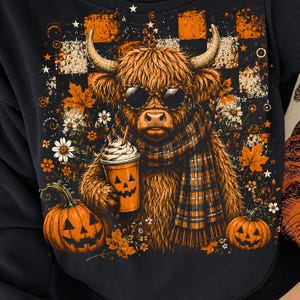 May include: A black sweatshirt featuring a graphic of a shaggy Highland cow wearing leopard-print sunglasses and a plaid scarf. The cow holds a pumpkin-faced cup with whipped cream, surrounded by orange pumpkins, autumn leaves, and flowers.