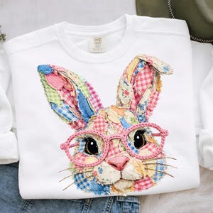 Puede incluir: Sudadera blanca con un diseño de conejo de retazos con gafas rosas. La cara y las orejas del conejo están hechas de parches de tela coloridos con patrones de cuadros y flores. El conejo tiene una nariz rosa y bigotes.