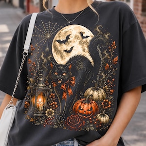 May include: A charcoal gray t-shirt featuring a graphic print of a black cat with a bow, a glowing lantern, pumpkins, a full moon, bats, and a spiderweb. The design incorporates orange and yellow autumn-themed floral accents and foliage.
