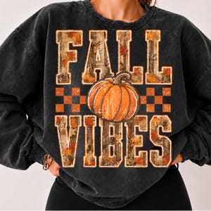 May include: A charcoal black crewneck sweatshirt featuring a graphic print with the text FALL VIBES in block letters. The letters are filled with a camouflage and autumn leaf pattern. A large orange pumpkin sits between the two words.