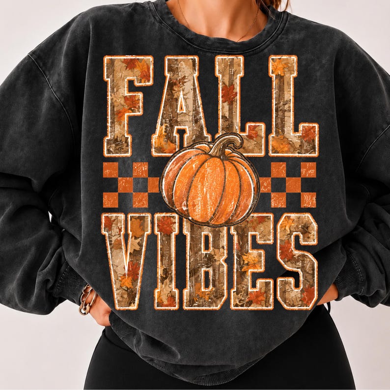 May include: A charcoal black crewneck sweatshirt featuring a graphic print with the text FALL VIBES in block letters. The letters are filled with a camouflage and autumn leaf pattern. A large orange pumpkin sits between the two words.