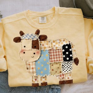 May include: A light yellow sweatshirt featuring a patchwork cow design. The cow is made of various fabric patterns, including polka dots, gingham, and solid colors. The sweatshirt has a crew neck and long sleeves.