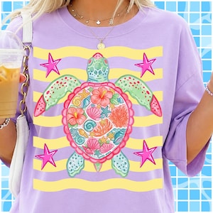 May include: A lilac-colored t-shirt featuring a sea turtle design. The turtle's shell is filled with seashells and flowers, with green and pink accents. Yellow horizontal stripes and pink stars complete the design.