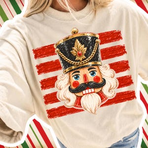 Puede incluir: Una sudadera de cuello redondo color crema con un estampado gráfico de una cabeza de cascanueces que lleva un sombrero negro con adornos dorados y una joya roja. El cascanueces tiene bigote negro, barba blanca y ojos azules sobre rayas rojas.