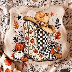 Könnte beinhalten: Ein beiges Sweatshirt mit einem Grafikdruck, der einen beigen Cowboyhut auf zwei Cowboystiefeln zeigt. Ein Stiefel hat ein Herbstblattmuster, der andere ein schwarz-weißes Schachbrettmuster. Orangefarbene und weiße Kürbisse sowie Herbstlaub umgeben sie.