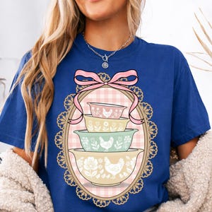 Puede incluir: Camiseta azul con un diseño de cuatro cuencos para mezclar en colores pastel, cada uno con motivos florales y de gallinas. Los cuencos están dentro de un marco ovalado con un fondo de cuadros rosas y un lazo rosa. El marco está rodeado por un borde de encaje decorativo.