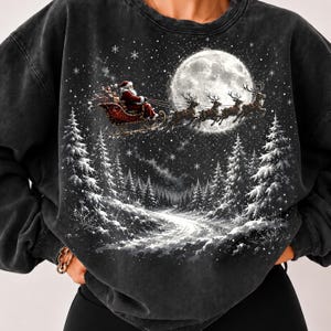 Op de afbeelding: Een antracietkleurige sweater met ronde hals en een grafische print van de Kerstman in een arrenslee getrokken door rendieren die voor een grote volle maan vliegen. De scène toont een besneeuwd boslandschap met dennenbomen en sneeuwvlokken.