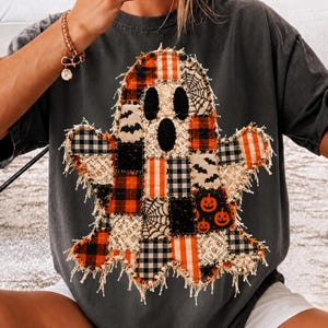 May include: A charcoal gray t-shirt featuring a large ghost-shaped graphic made of various fabric patches. The patchwork ghost includes orange and black plaid, white lace, black bat prints, spider webs, and orange jack-o'-lantern patterns.