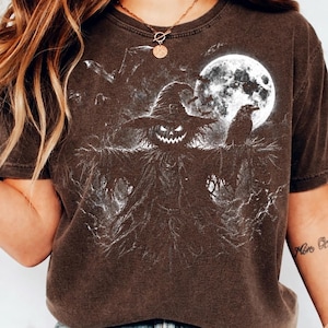 May include: A dark brown short-sleeve t-shirt featuring a white graphic print of a scarecrow with a jack-o'-lantern head wearing a wide-brimmed hat, standing beneath a full moon with a raven perched on its arm and bats flying in the background.